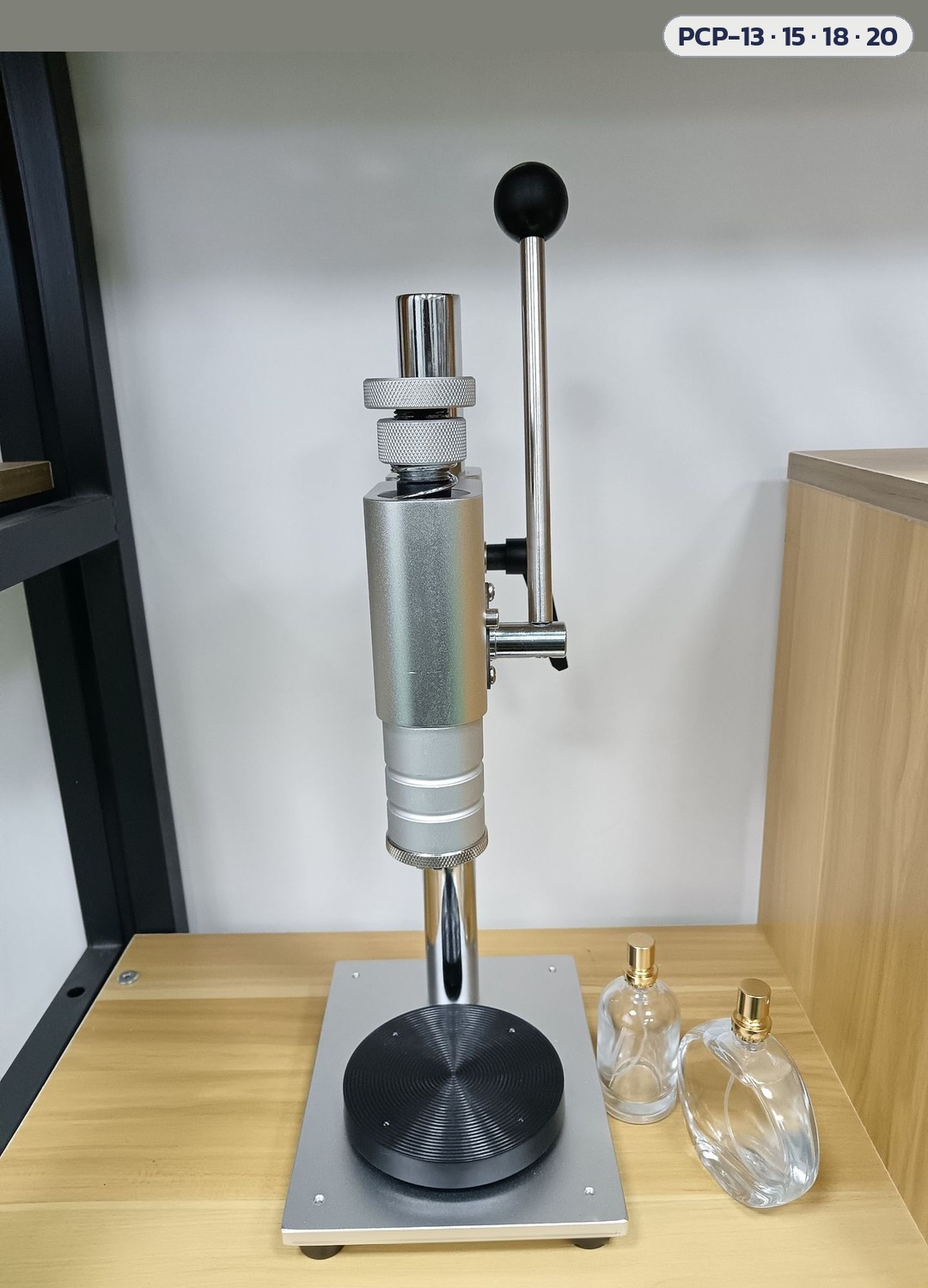 เครื่องย้ำปลอกฝา/กดหัวสเปรย์น้ำหอม มือโยก PCP-13 / 15 / 18 / 20 – ภาพถ่ายเครื่องด้านข้างบนโต๊ะ แท่นวางสีดำ เห็นแกนคันโยกและหัวกด (ขวดน้ำหอมในภาพเป็นของประกอบฉาก ไม่รวมในชุดเครื่อง)